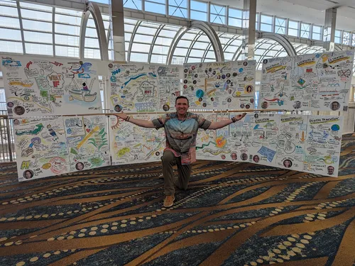 Graphic Recorder Matt Orley kneeling with his hands raised in front of a subset of drawings from a pharmaceutical association conference.