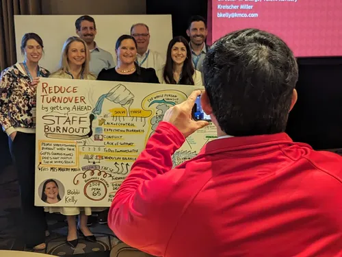 Photo of a photographer taking a picture of a conference session speaker and her cohort.  The speaker is holding up the graphic recording/illustration.