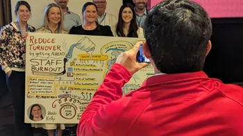 Photo of a photographer taking a picture of a conference session speaker and her cohort.  The speaker is holding up the graphic recording/illustration.