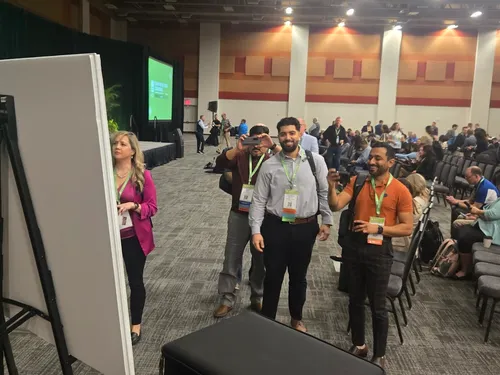 Event attendees eyeing a graphic recording created during a  keynote address.  Photograph taken from behind the panel, no art is visible, but their positive reactions are visible.