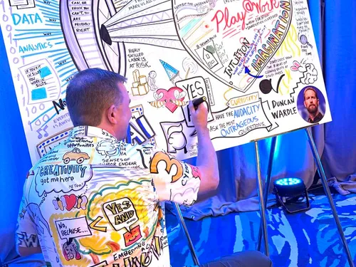 Matt Orley kneeling in front of a graphic recording while holding a marker against the board.  Matt is wearing a shirt with a previously drawn illustration.  It looks like camouflage!